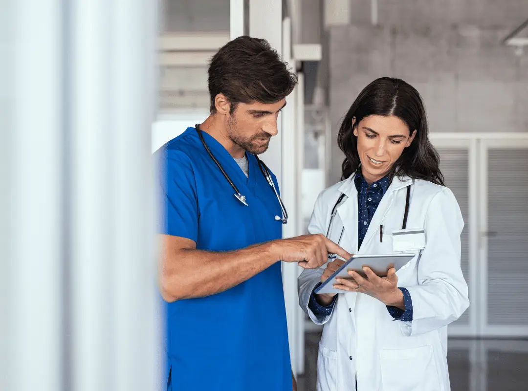 Two healthcare professionals, one in blue scrubs and the other in a white coat, discuss information on a tablet. They appear focused and collaborative.