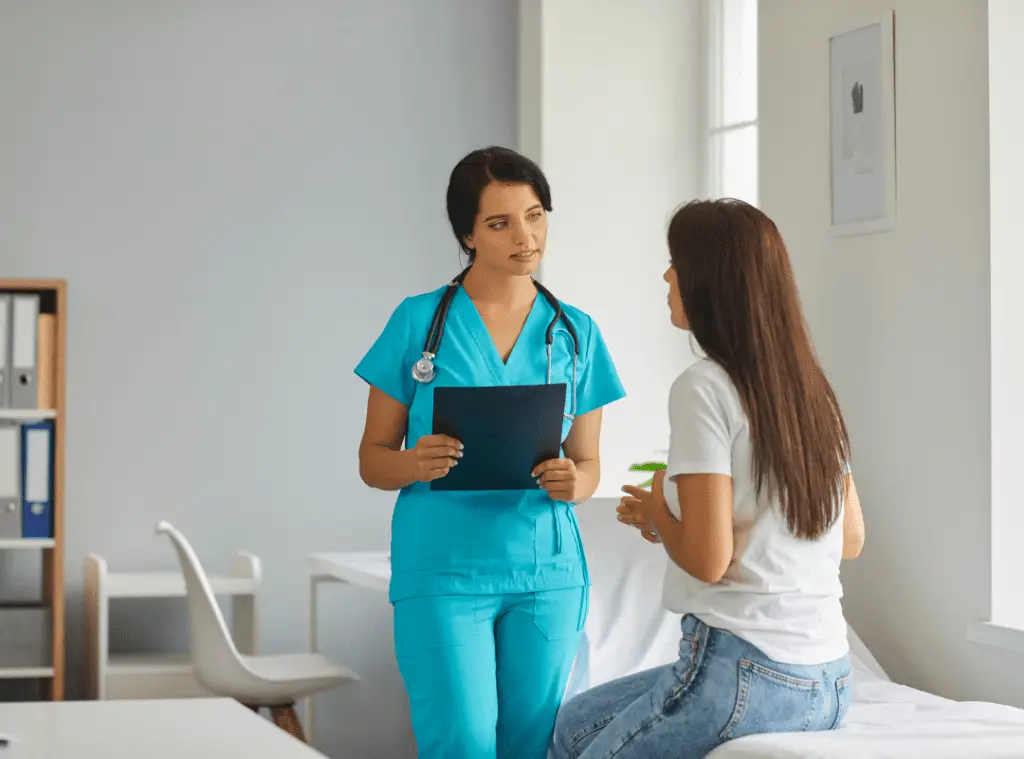 A nurse in blue scrubs holds a clipboard, talking to a seated woman in casual clothes in a bright, tidy medical office, conveying a calm and caring atmosphere.