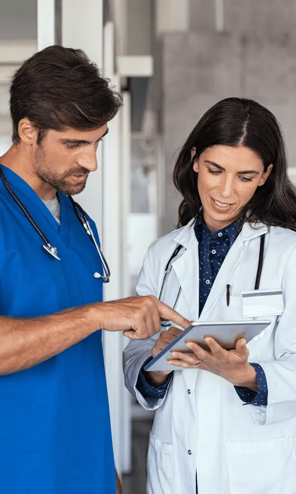 Two healthcare professionals, one in blue scrubs and the other in a white coat, discuss information on a tablet. They appear focused and collaborative.