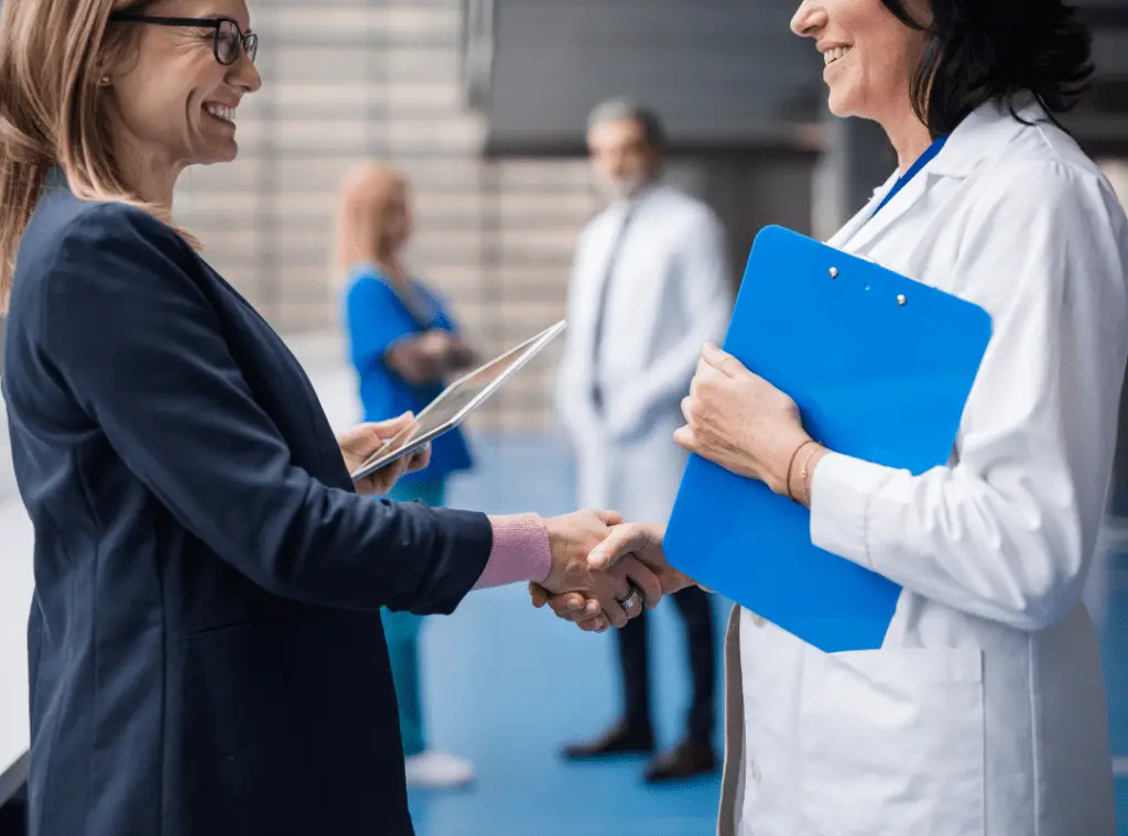 Two women, a healthcare worker and a business professional, smile and shake hands in a medical setting. Two more healthcare professionals are blurred in the background.