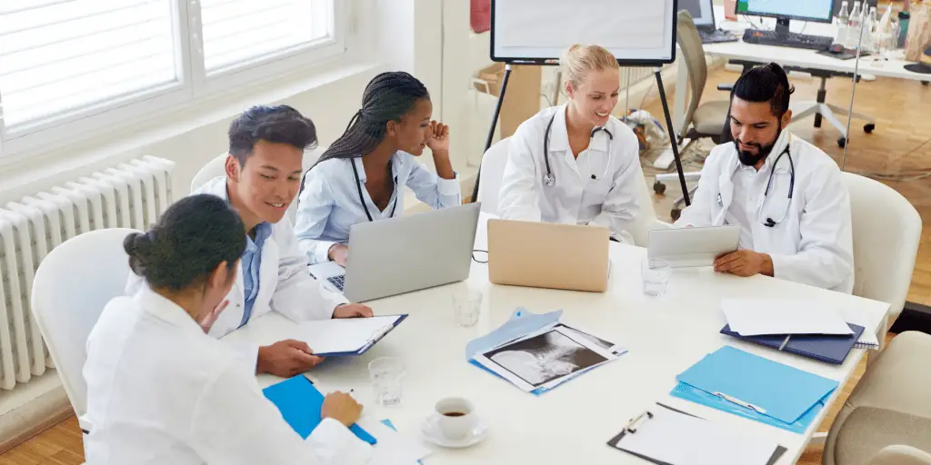 A diverse group of five medical professionals in white coats sit around a table, collaborating with laptops and papers, creating a focused and cooperative atmosphere.