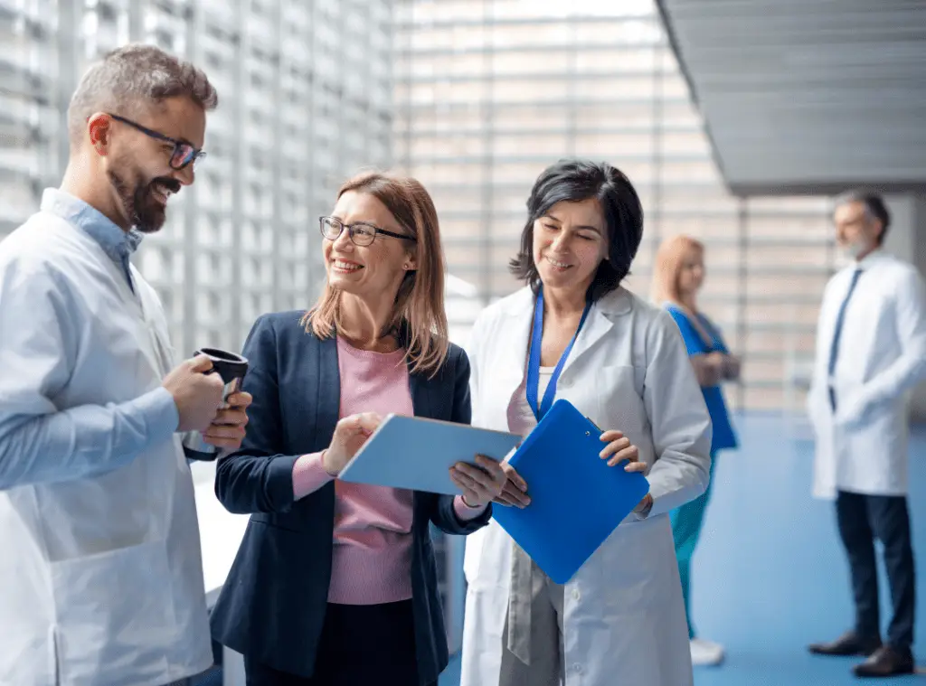 A group of medical professionals converse in a bright hallway. Three doctors in white coats and a woman in a suit smile, holding a tablet and clipboard.