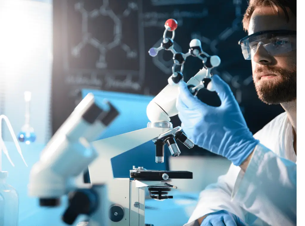 A scientist in goggles and gloves examines a molecular model beside a microscope. The background shows a chalkboard with chemical diagrams.