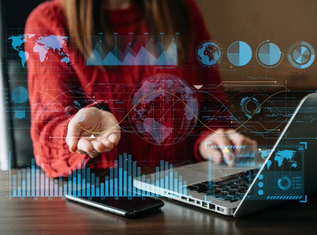 A person in a red sweater sits at a desk with a laptop. Holographic charts and a globe appear above an open hand, suggesting data analysis and technology.